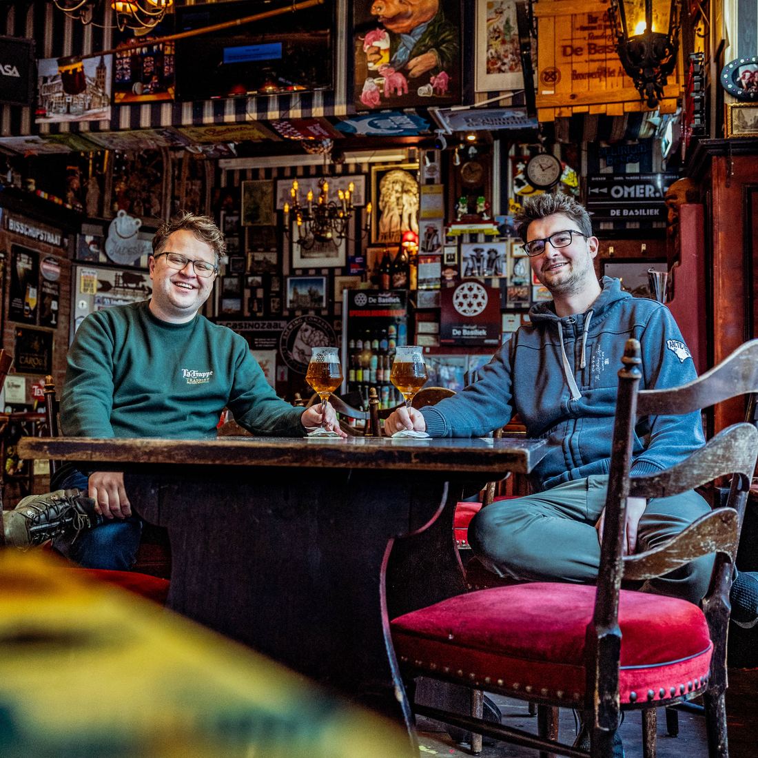 Twee mannen aan een houten tafel in een bruin biercafé vol bordjes en schilderijen