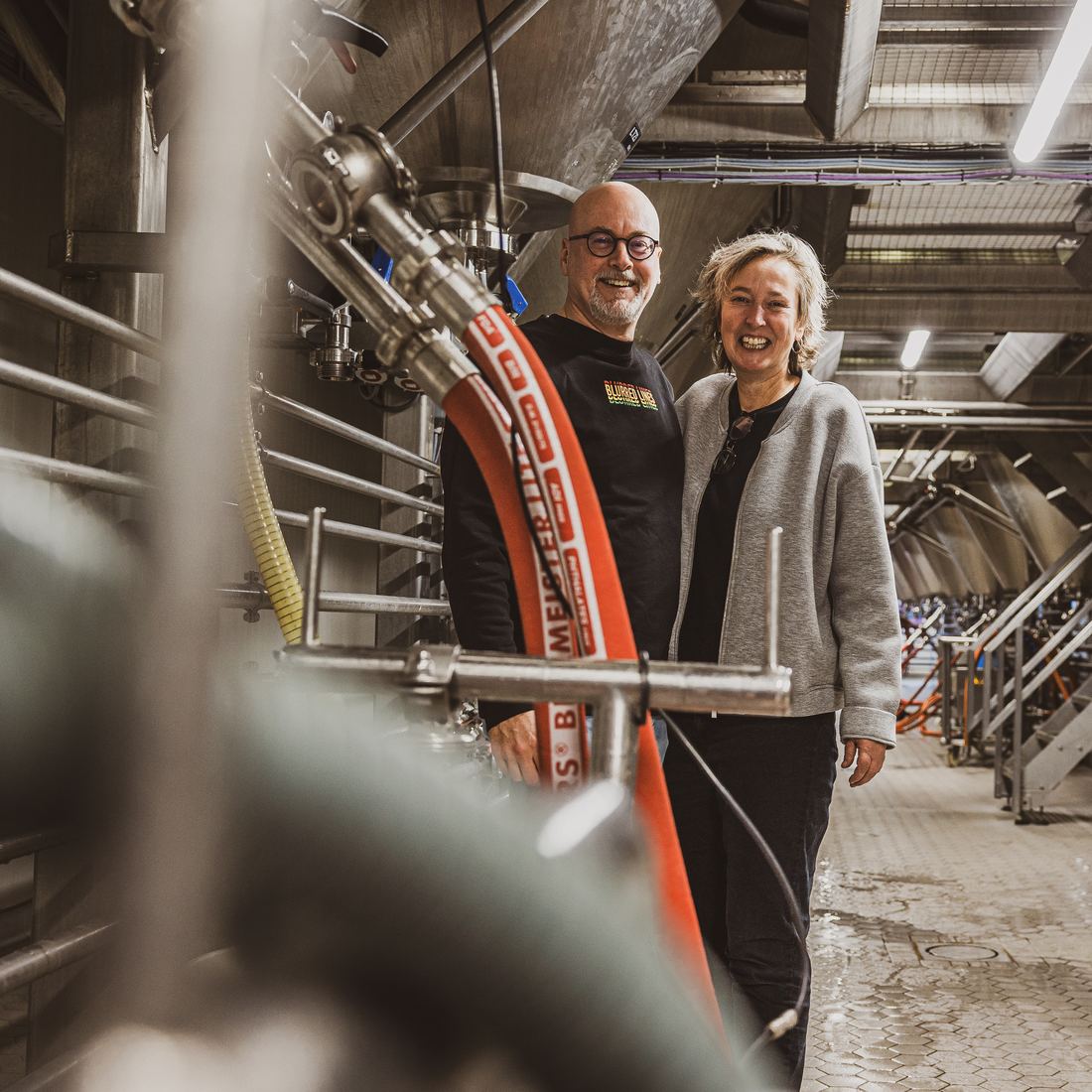 Michel Ordeman en Lydian Zoetman lachend tussen de leidingen en tanks van de brouwerij