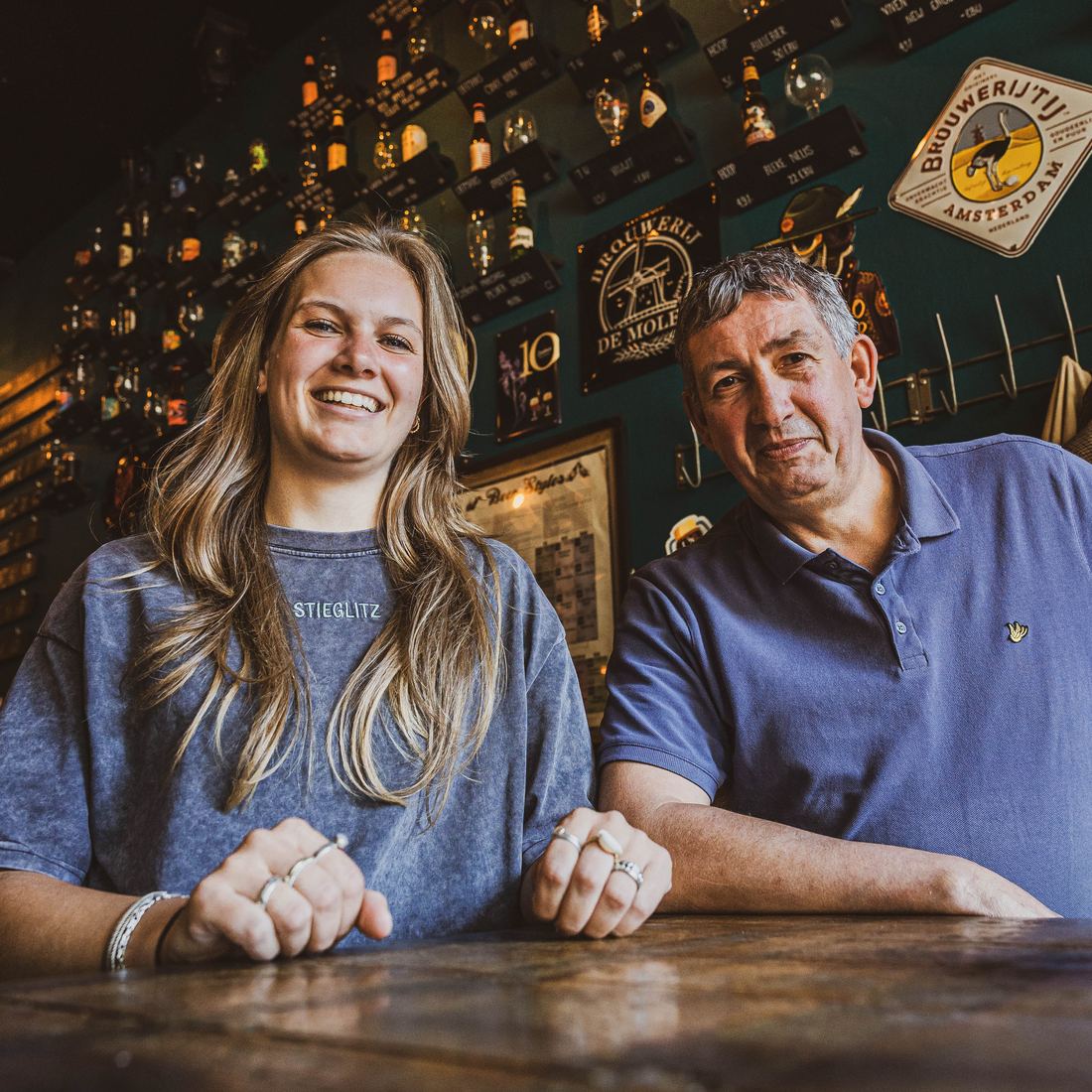 Tess en Hans Huberts aan de bar van Café Persee, met bierflessen en borden aan de muur